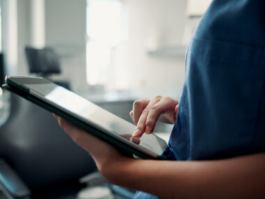 A healthcare professional, shown from the chest to the waist, uses a tablet.