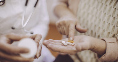 A patient holds out a hand filled with oral medications while a doctor examines a prescription bottle.