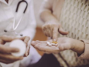 A patient holds out a hand filled with oral medications while a doctor examines a prescription bottle.