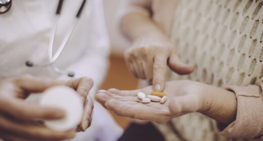 A patient holds out a hand filled with oral medications while a doctor examines a prescription bottle.