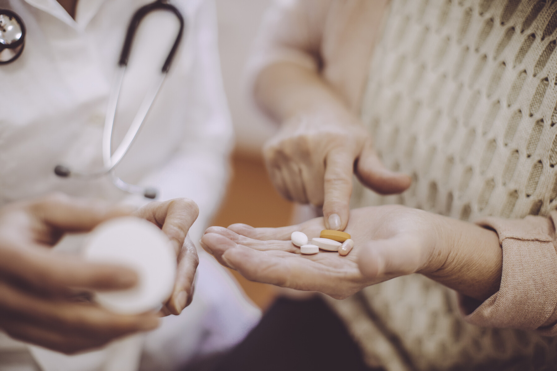 A patient holds out a hand filled with oral medications while a doctor examines a prescription bottle.